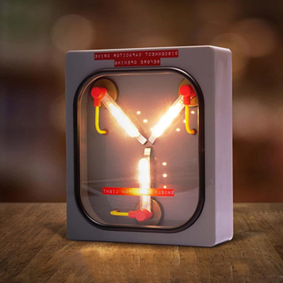 A mood lamp in the shape of the Flux Capacitor from the movie Back to the Future is displayed shining bright on a wooden desk glowing with yellow moody colors illuminating the red and grey interior of the flux capacitor.