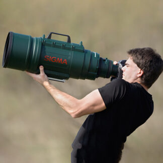 A person with a black t shirt is taking a photo with his huge camera lens attached to his camera. The lens is at the size of a bazooka and it's color is green.