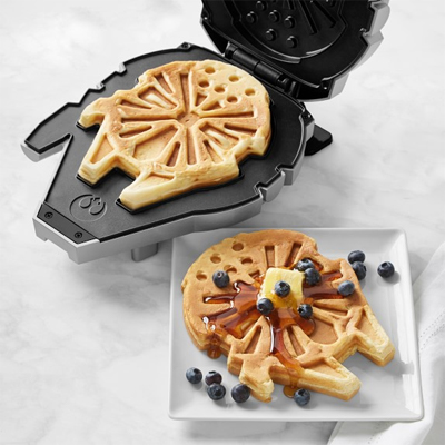 A waffle maker in the shape of the Millennium Falcon spaceship from Star Wars movie series is displayed with a waffle next to it in the shape of Millennium Falcon decorated with berries and butter on top of it.