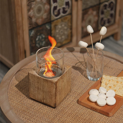 A miniature tabletop fireplace is displayed burning inside a cylindrical glass which is seated on an authentic wooden cube. There are marshmallows impaled to little sticks ready to be cooked on the fireplace.