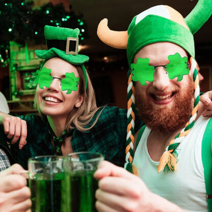 A man with a green and white viking hat and a girl with a green leprechaun hat are wearing green glasses in the shape of shamrock celebrating St. Patrick's Day having fun and drinking green beer.