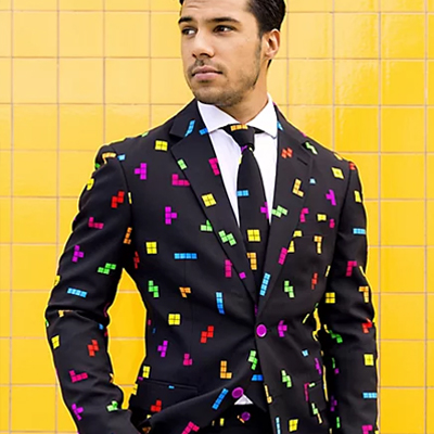 A man is displayed in front of an orange background wearing a Tetris patterned suit of complementing jacket and tie. The suit pattern is consisting of colorful blocks from the Tetris game on a black fabric.