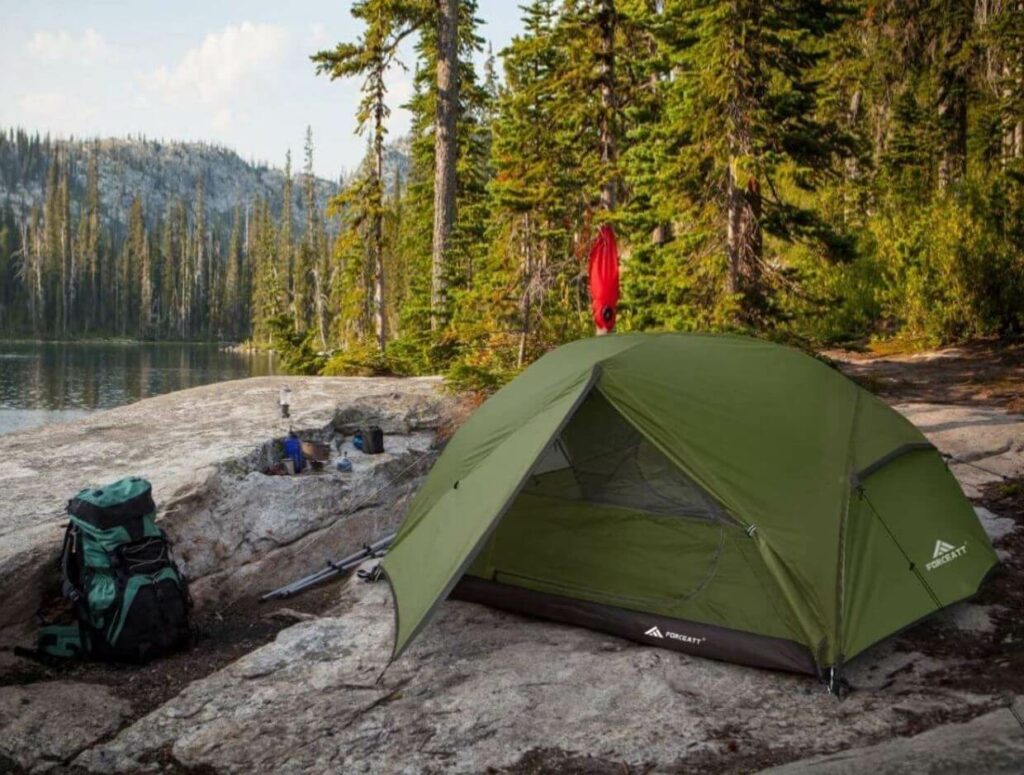 A green tent with a waterproof ceiling is installed on a rocky surface next to a river right next to the forest.